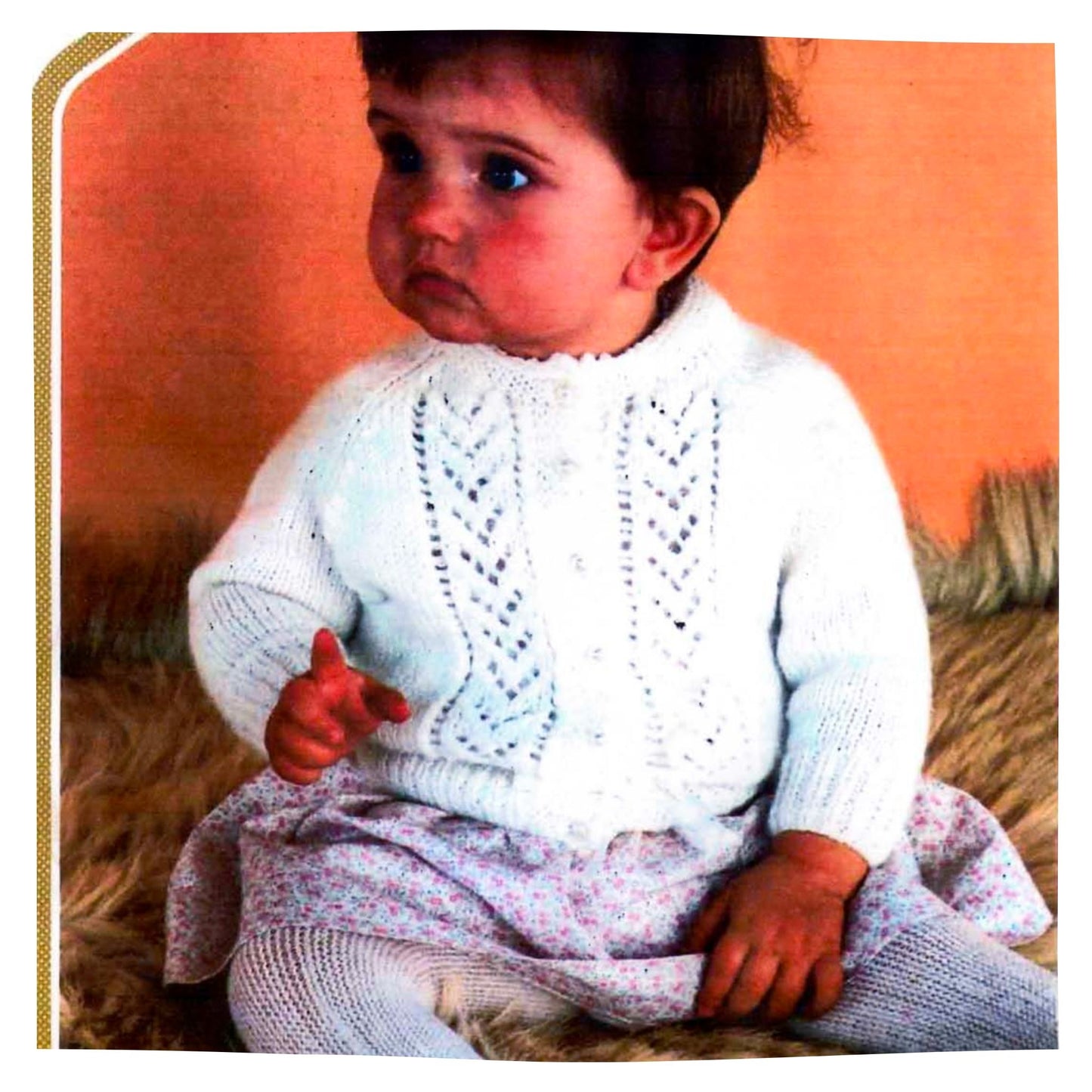 Baby wearing a white knitted sweater with a textured pattern against an orange background - this is a free vintage baby cardigan knitting pattern 
